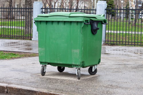 A large green wheeled rubbish bin made of hard plastic with a textured surface and a slightly domed lid, situated on a paved sidewalk in an outdoor setting. The bin's black handle is visible on the front, and the lid is closed, with the bin positioned on a concrete surface. In the background, there is a black metal fence with vertical bars, behind which there are trees with bare branches and a grassy area, suggesting a park or communal space. The scene appears to be during overcast weather, with diffused lighting. This image relates to waste collection and rubbish removal services, characteristic of local waste management in Maida Vale.