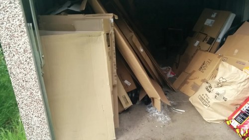 An outdoor storage area or garage space showing a variety of discarded items and packaging materials. In the foreground, a large, cream-colored wooden or particleboard cabinet or shelving unit leans against a gravel or concrete surface. To the right, there is a pile of flattened cardboard boxes, some with printed labels and branding visible, along with crumpled brown paper and plastic wrapping. The background features a dark interior space, possibly a garage or basement, with additional cardboard boxes and miscellaneous rubbish stacked or stored. The environment suggests a clearing or collection of waste materials, consistent with rubbish removal services in the Maida Vale area, as provided by Commercial Waste Removal Maida Vale. The scene is lit with natural or ambient light, with a subtle contrast between the cluttered foreground and the darker indoor backdrop.
