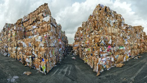 Two large, densely packed stacks of compacted cardboard and paper waste are positioned on either side of a gravel driveway or lot, with clear textures of corrugated cardboard and various paper materials visible. The waste appears to be organized into rectangular bales, with visible ripples and folds in the cardboard surfaces, and some printed materials and labels can be discerned among the paper. The stacks extend upwards and occupy most of the horizon, creating a high-walled enclosure with a narrow passage between them. The sky overhead is cloudy, casting diffuse natural light over the scene, emphasizing the rough textures and earthy tones of the waste. In the background, a small section of a dark vehicle or machinery may be partially visible behind the waste piles. This image relates to commercial rubbish management and waste removal services in Maida Vale, London, as represented by the context of the scene, aligning with services offered by Commercial Waste Removal Maida Vale.