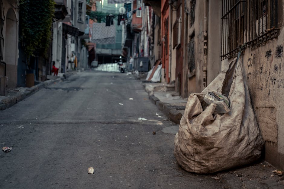 An outdoor scene showing a large heap of mixed domestic and commercial waste overflowing from several black, grey, and red wheelie bins placed on the pavement near a car park. The waste includes crump