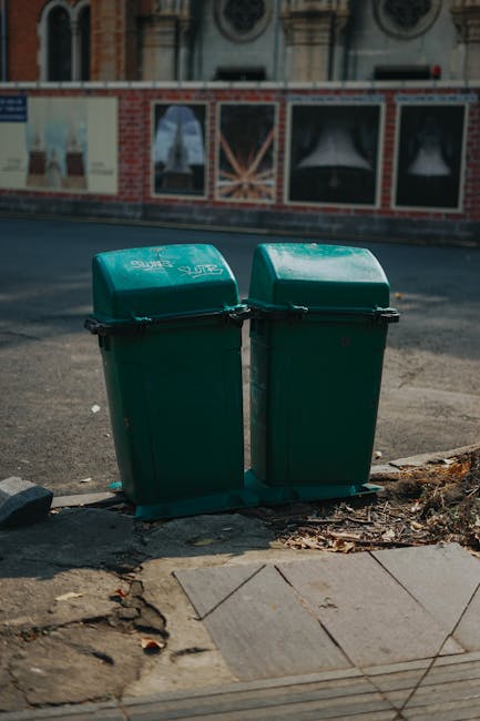 Two green wheeled rubbish bins made of plastic with slightly curved, domed lids are positioned side by side on a concrete pavement at the edge of a street, near a curb. The bins have black handles and
