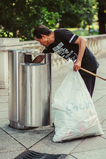 Two green wheeled rubbish bins made of plastic with slightly curved, domed lids are positioned side by side on a concrete pavement at the edge of a street, near a curb. The bins have black handles and